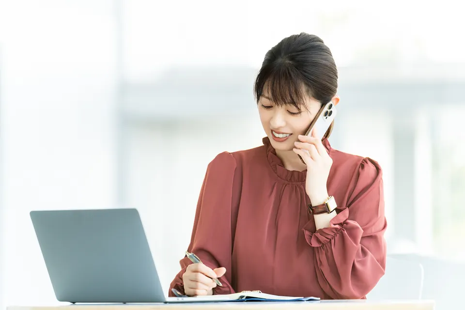 ノートパソコンの前で電話を受けながらメモを取る女性