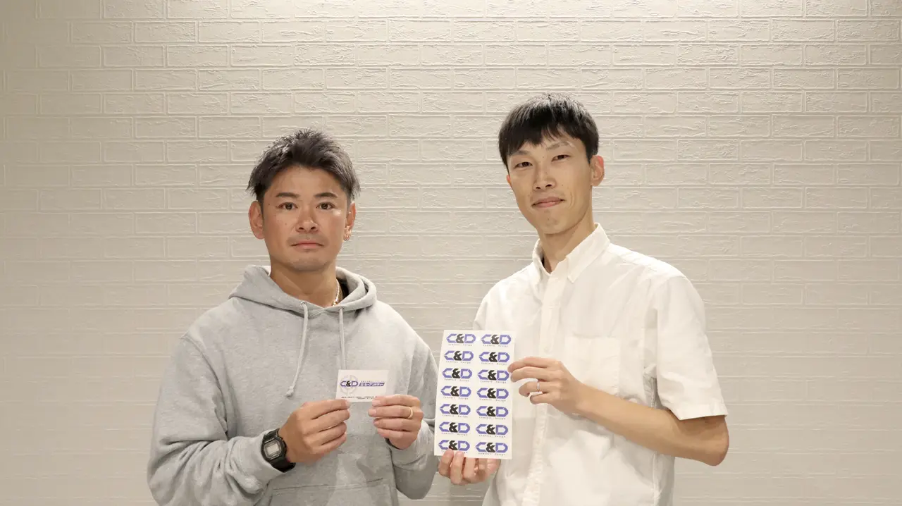 C&Dの松田さまと笠原 友生。シールのシートと名刺を手に並ぶ様子